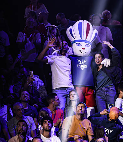 A group of people are sitting in the stands, watching a mascot perform. The mascot is a large, costumed figure of a person wearing a white shirt and a blue hat. The crowd is engaged and enjoying the performance. AI generated content