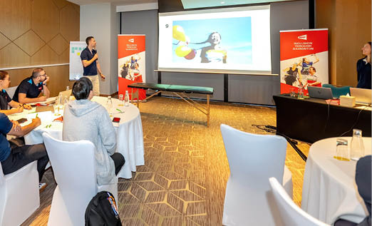 A group of people are sitting in a conference room, watching a presentation on a large screen. There are several chairs and dining tables in the room, and a few people are wearing ties. The attendees are engaged in the presentation, which is displayed on a large TV screen. AI generated content