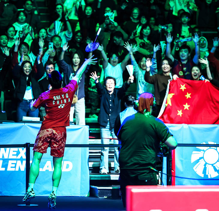 A man in a red shirt is holding a tennis racket, surrounded by a crowd of people. The crowd is waving and cheering, with some of them holding up their cell phones. A woman in the crowd is holding a Chinese flag. AI generated content